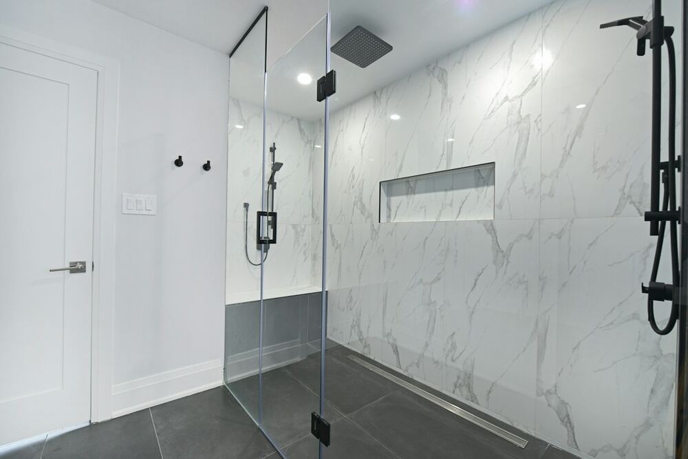 Puerta de ducha de vidrio sin marco con herrajes negros en un baño con paredes de mármol